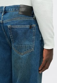 Pogled od blizu na osebo, ki nosi modre AllSaints kavbojke, kjer je vidna zadnja žep, usnjena oznaka blagovne znamke in roka, ki počiva blizu stegna.