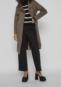 Long manteau en daim beige avec un col cranté, pull rayé noir en dessous, pantalons larges noirs, et sandales noires à talons épais.
