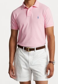 Rosa polo skjorta med korta ärmar, prydd med ett blått broderat emblem. I kombination med vita shorts och ett brunt bälte, samt en klocka på handleden.