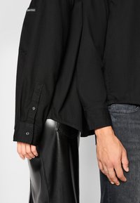 Črna dolgosleeve majica iz gladkega blaga, z gumbi na manšeti in subtilnim logotipom. V kombinaciji s sijočimi črnimi usnjenimi hlačami in zbledelimi kavbojkami.