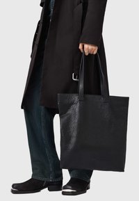 Zwarte leren tote bag met een getextureerde afwerking en lange handvatten. De tas is vierkant van vorm en wordt gedragen door een persoon in donkere kleding.