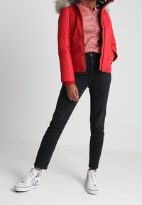 Rød puffet jakke med en faux fur hætte, parret med en lys pink skjorte med knapper og sorte slim-fit jeans. Hvide sneakers med mønstrede detaljer.