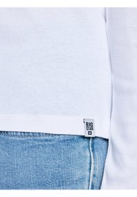 Balta krekla ar garām piedurknēm ielikta gaiši zilu džinsu biksēs, uz krekla apakšējās sānu šuves redzama maza "BIG STAR" zīmola etiķete.