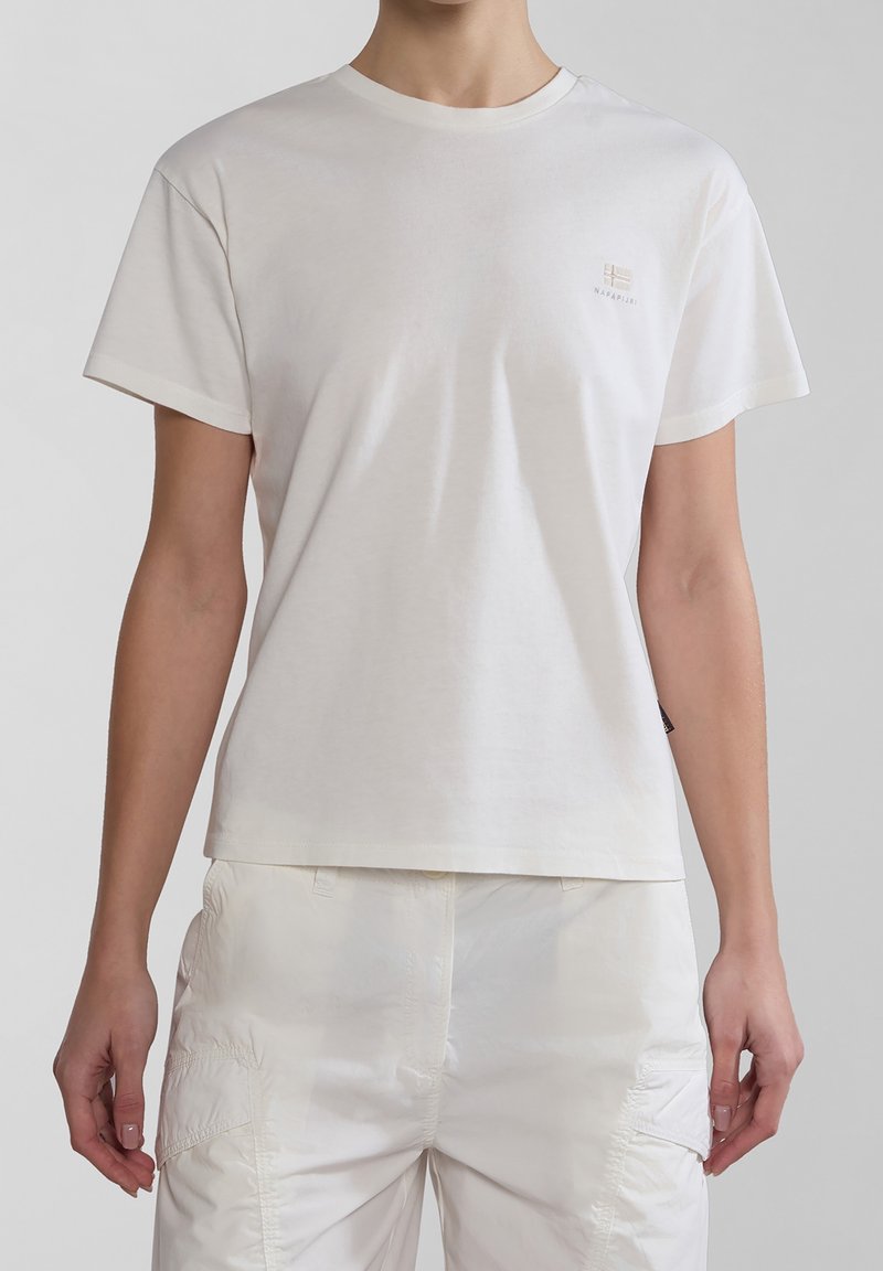 Personne portant un T-shirt blanc uni à manches courtes avec un petit logo sur le côté gauche de la poitrine et un pantalon de couleur claire avec des poches latérales.