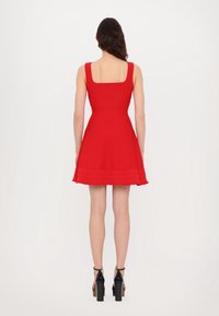 Femme debout de dos, portant une robe rouge évasée sans manches et des chaussures à plateformes noires à talons hauts, sur fond blanc uni.