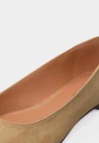 Μπεζ σουέτ επίπεδο παπούτσι με ελαφρώς μυτερή μύτη και μαλακή ταμπά εσωτερική επένδυση, εμφανής πάνω σε λευκό φόντο.