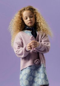 Cardigan en tricot rose clair avec des détails en rose, associé à une jupe en denim bleu à motifs. Le modèle porte une écharpe bleu foncé, sur un fond violet.