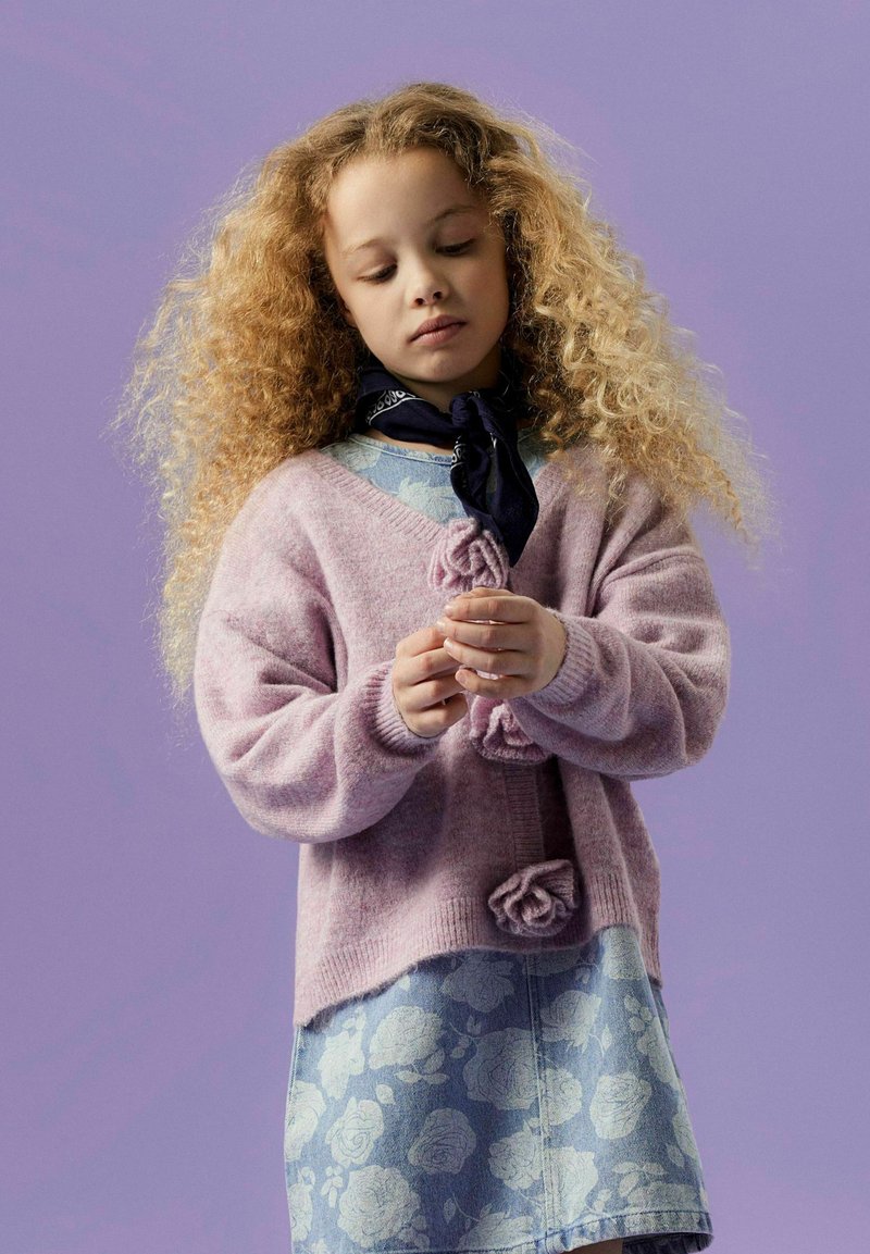 Cardigan en tricot rose clair avec des détails en rose, associé à une jupe en denim bleu à motifs. Le modèle porte une écharpe bleu foncé, sur un fond violet.