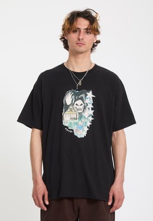 Jeune homme aux cheveux ondulés portant un t-shirt noir surdimensionné orné d'un graphisme représentant une maison, une figure au visage de crâne, des étoiles et une petite personne faisant signe de la main.