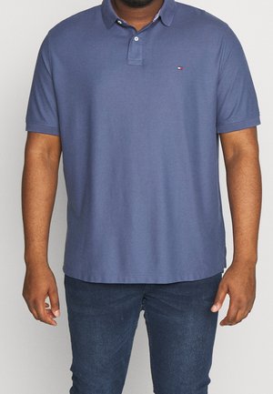 Mann trägt ein blaues Poloshirt mit kurzen Ärmeln, geknöpftem Kragen und kleinem rot-weiß-blauem Logo auf der Brust, kombiniert mit dunkelblauen Jeans.