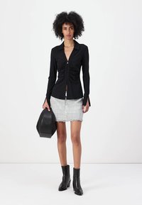 Blusa nera a maniche lunghe con arricciatura frontale e chiusura con zip, abbinata a una mini gonna grigia con texture e stivali neri alla caviglia, mentre tiene una borsa nera.