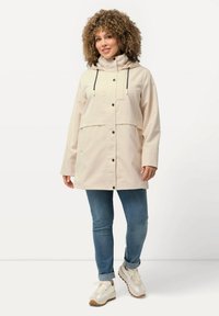 Giacca impermeabile beige con cappuccio, coulisse e chiusura frontale a bottoni. Presenta grandi tasche frontali e un design dal taglio rilassato.