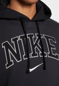Czarna bluza z kapturem z dużym szarym logo „NIKE”. Posiada białe przeszycia i sznurki. Materiał bawełniany o gładkiej fakturze.