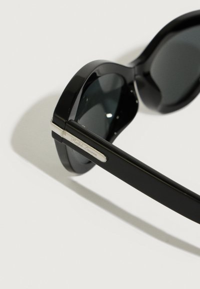 Μαύρα γυαλιά ηλίου με σκούρους φακούς, που διαθέτουν φαρδύ σκελετό, γυαλιστερή επιφάνεια και ασημί αξεσουάρ. Χαραγμένο λογότυπο στο πλάι.