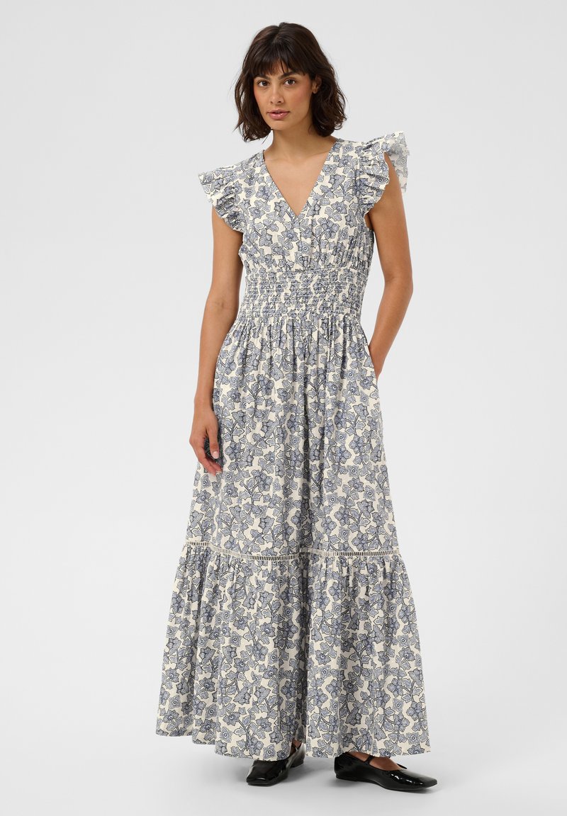 Femme portant une robe fleurie sans manches, longue jusqu'aux chevilles, avec des épaules à volants et des chaussures plates noires, debout devant un fond uni.