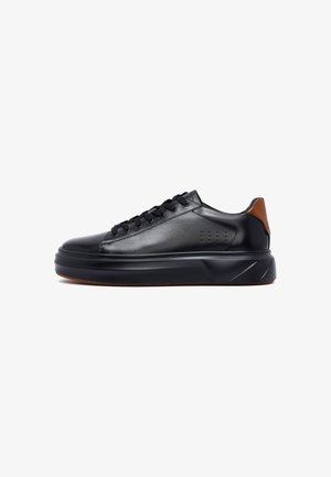 Zwarte leren sneaker met dikke zool, ronde neus, zwarte veters en bruine hieltab, weergegeven in profiel op een witte achtergrond.