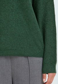 Maglione lavorato a maglia di color verde scuro con polsini a coste, indossato sopra pantaloni di tessuto grigio. Primo piano che mostra i dettagli della tessitura e la lunghezza delle maniche.
