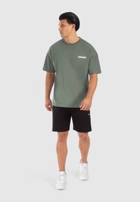 Camiseta de algodón verde oliva de manga corta y cuello redondo, con un logotipo blanco de "SMILODOX" en el lado izquierdo del pecho. Combinada con pantalones cortos negros.