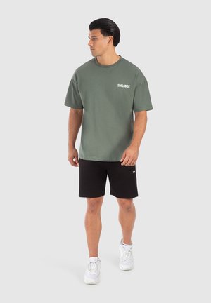 Olijfgroen katoenen t-shirt met korte mouwen en ronde hals, met een witte "SMILODOX" logo op de linkerborst. Gecombineerd met zwarte shorts.