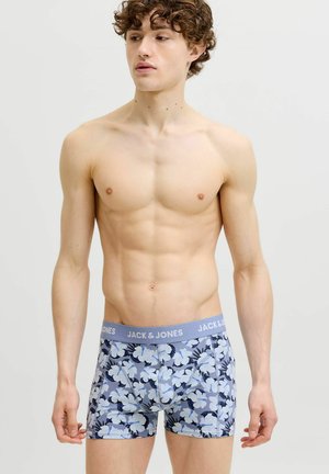 Jeune homme aux cheveux bouclés portant un boxer bleu à motif floral avec une ceinture « JACK & JONES », debout devant un fond uni.