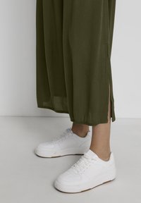 Baskets blanches en cuir lisse et texturé, avec des lacets plats et une semelle en caoutchouc, associées à un vêtement fluide d'un vert olive, longue jusqu'au sol, avec des fentes sur les côtés.