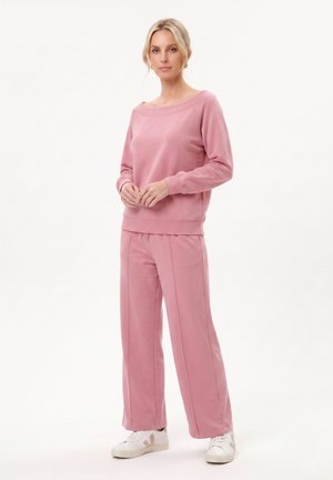 Kvinde, der står iført en pink bluse med lange ærmer og matchende vidde bukser med hvide sneakers på ensfarvet baggrund.