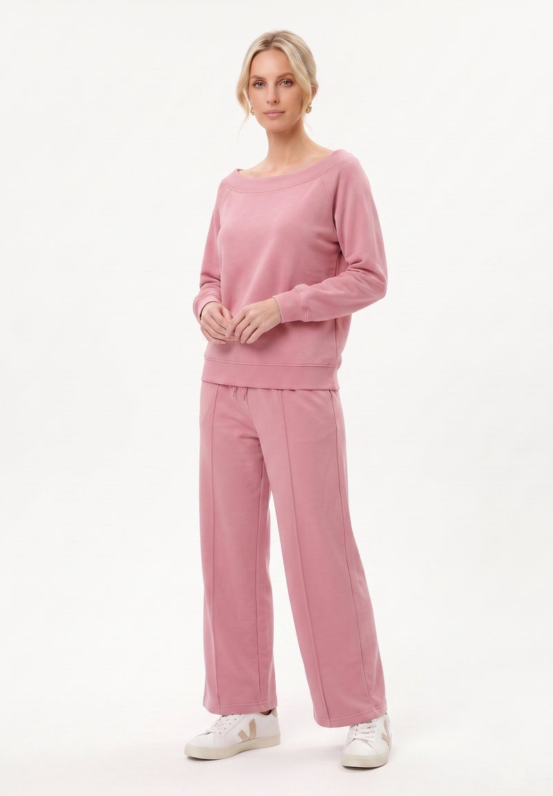Femme debout portant un haut à manches longues rose et un pantalon large assorti avec des baskets blanches sur un fond uni.