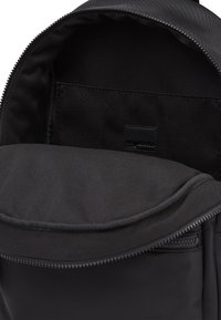 Μαύρη τσάντα πλάτης με εσωτερικό από λείο, ματ ύφασμα. Περιλαμβάνει μια κύρια θήκη και μια μικρή τσέπη με φερμουάρ. Χωρίς ορατά μοτίβα ή υλικά.