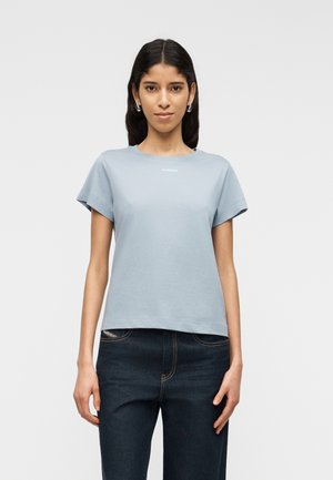 Kobieta o ciemnych włosach, ubrana w jasnoniebieski krótki rękaw t-shirt i ciemnoniebieskie dżinsy, stoi na tle jednolitego jasnego tła, zwrócona przodem.