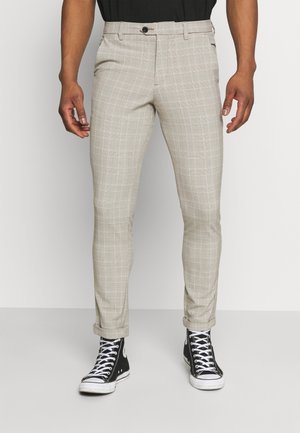 Man wearing slim-fit beige plaid trousers with cuffed hems and black high-top sneakers, standing against a plain background.