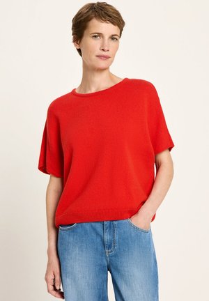 Frau mit kurzen braunen Haaren, die einen lockeren roten kurzärmeligen Pullover und blaue Jeans trägt, eine Hand in der Tasche, neutraler Hintergrund.