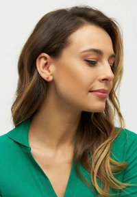 Pendiente de tachuela en forma de corazón rojo en una modelo con pelo ondulado de longitud media. La modelo lleva una blusa verde con collar, lo que resalta el detalle del pendiente.