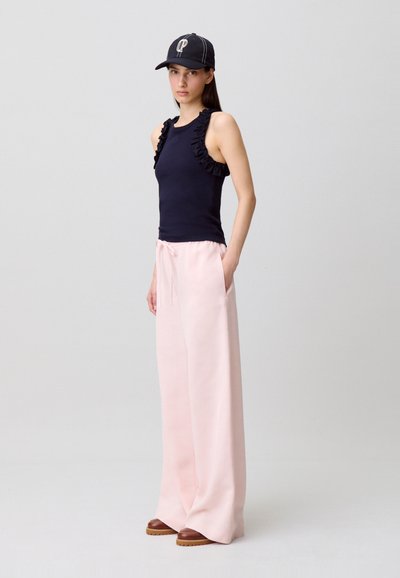 Femme portant un haut sans manches noir à volants, un pantalon large rose clair, des chaussures marron et une casquette noire, debout devant un fond uni.