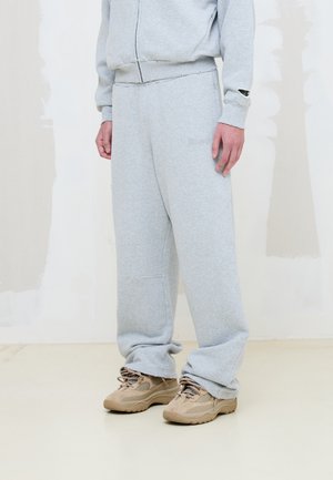 Persona que lleva pantalones deportivos gris claro y sudadera a juego con zapatillas beige, de pie sobre un suelo claro contra una pared pálida.