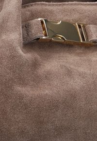 Bruine suède tas met een soepele textuur en een metallic gouden sluiting als detail op de riem. De algehele vorm is zacht en rond.
