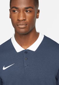 Marineblaues Poloshirt mit weißem Kragen, einer Knopfleiste mit drei Knöpfen und dezentem Nike-Swoosh-Logo auf der linken Seite. Material aus Baumwollmischung, glatte Textur.