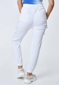 Witte joggingbroek van rekbaar materiaal, met twee zijzakken en elastische boorden. Gepaard met lichtgekleurde sportschoenen.