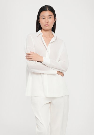 Camicia bianca con bottoni, maniche lunghe, vestibilità rilassata, texture morbida e colletto classico. Indossata con pantaloni bianchi abbinati.