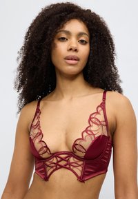 Σουτιέν σε μπουρντόρ με διάφανα τούλινα στοιχεία, κυματιστά άκρα και σχέδιο σταυρού στο μπροστινό μέρος. Λείες σατινέ θήκες και ρυθμιζόμενες τιράντες.