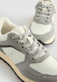 Graue Wildleder- und cremefarbene Leder-Sneaker mit strukturierten Akzenten, flachen Schnürsenkeln, gepolstertem Kragen und einer Gummisohle in neutralem Farbschema.