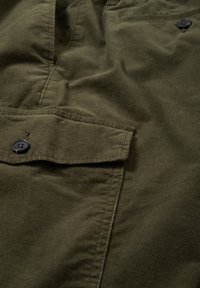 Gros plan sur un tissu en velours côtelé vert olive avec une poche à rabat boutonnée et des détails de couture visibles.