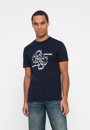 Joven con una camiseta gráfica azul marino con letras blancas y negras y jeans desgastados, de pie frente a un fondo blanco liso.