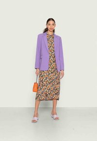 Blazer lavanda sobre um vestido floral escuro com flores laranjas e amarelas, combinado com sandálias de salto bloco roxas e uma bolsa laranja.