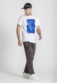 T-shirt bianco in cotone con una grande grafica di un orsetto gommoso blu, abbinata a pantaloni marroni a sigaretta e sneaker bianche, indossata con un cappellino nero.