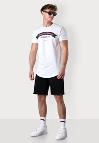 White cotton t-shirt with a black and pink graphic design reading "Summer Romance Marbella," paired with black denim shorts and white sneakers.