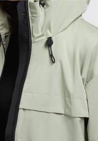 Jasnozielona kurtka z suwakiem, przednią kieszenią i regulowanym kapturem, z czarnymi detalami przy zamku i kołnierzu. Miękka w dotyku.