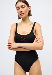 Μαύρο μαγιό με στρογγυλή λαιμόκοψη, φαρδιές τιράντες και ψηλές περικοπές στα πόδια. Διακοσμημένο με κόκκινη καρδιά και κείμενο "HUGO" στο μπροστινό μέρος.