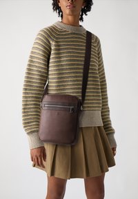 Ruda odinė crossbody krepšys su priekine užtrauktuko kišenę, lygiu paviršiumi ir reguliuojama dirželiu, dėvima virš dryžuoto megztinio ir klostuotos sijono.