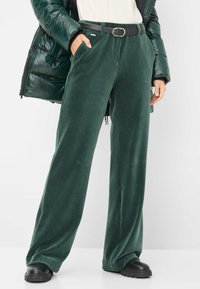Grüne Corduroy-Hose mit weitem Bein und seitlichen Taschen, kombiniert mit einem schwarzen Gürtel und klobigen schwarzen Schuhen; getragen mit einer glänzenden dunkelgrünen Daunenjacke.
