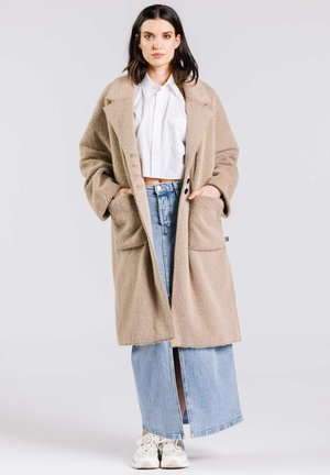 Manteau en laine beige surdimensionné avec revers, deux poches avant, boutons double boutonnage. Porté sur une jupe en denim bleu clair et des baskets blanches.